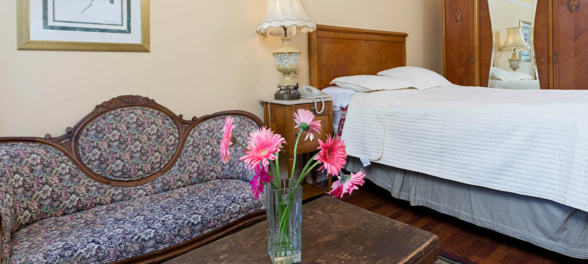 In a yellow guestroom a white bed rests on the wall acrosas from a purple floral couch