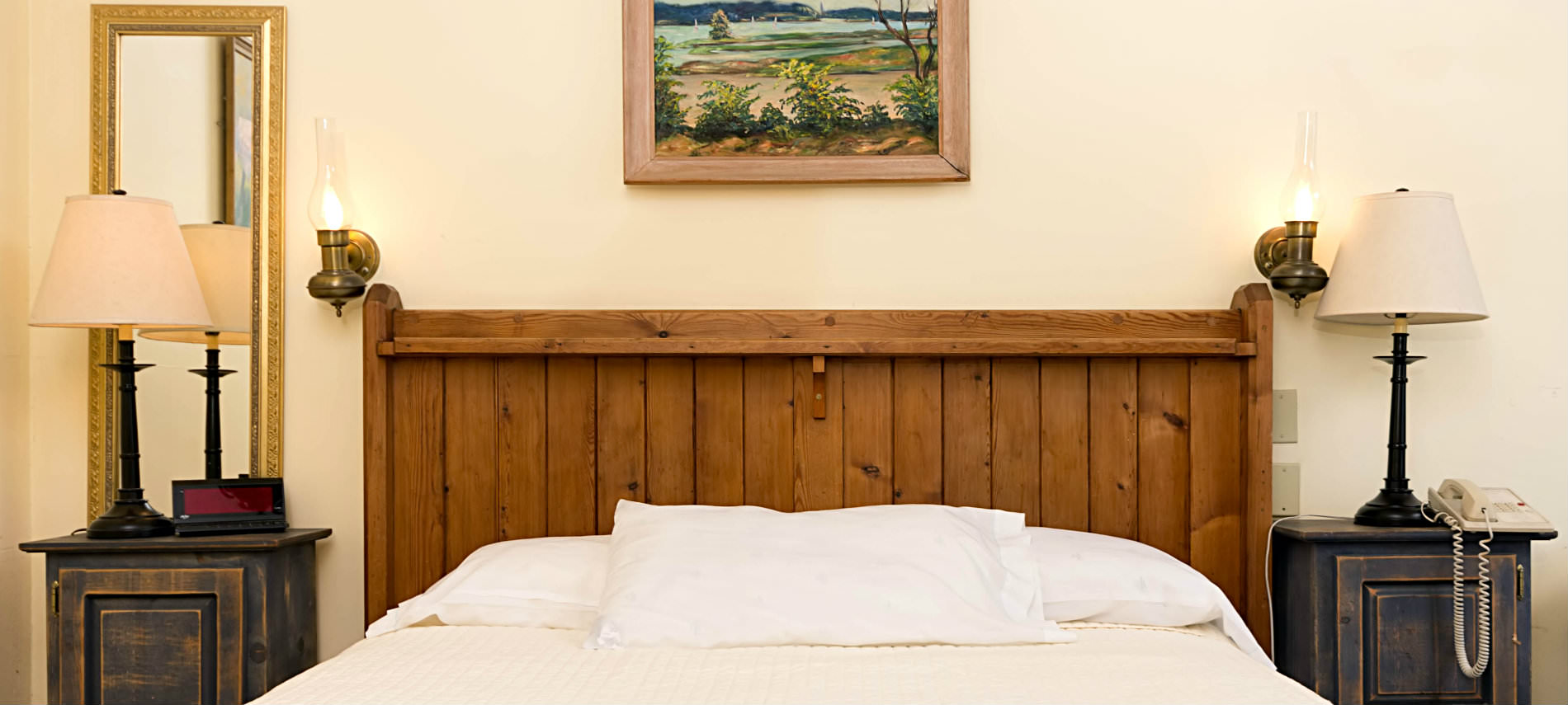 A whte bed with a pillow and a brown wooden headboard sits between two small tables with lamps