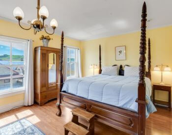 A bedroom with yellow walls, hardwood floors, an antique four poster bed with wood steps at the foot of the bed, wood nightstands with lamps, and an antique mirrored dresser in between two windows.