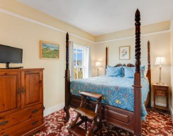 A bedroom with red and peach tapestry carpet, a wood four poster bed, a dresser in between two windows, and a sofa and coffee table along one wall.