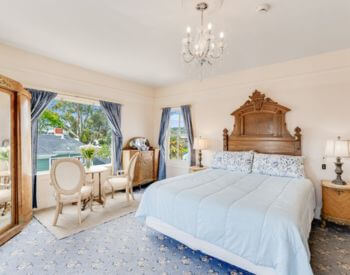 carpeted peach painted room with king bed and oak headboard, window with table and cushioned chairs, stands with lamps
