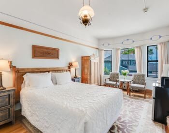 A bedroom with wood floors, light blue walls, a bed with blue and white bedding, nightstands with lamps on either side of the bed, and a bay window with a sitting area of 2 chairs and a small table.