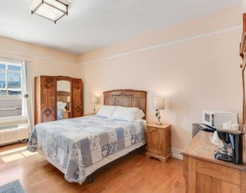 A bedroom with wood floors, peach colored walls, a bed with white bedding, a long sofa on the opposite wall, and a mirrored dresser and mini fridge and microwave along another wall.