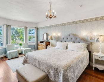 a bedroom with hardwood floors, light blue tapestry wallpaper, a bed with white and blue bedding, an antique mirrored dresser, a footstool at the foot of the bed, and a sitting area in front of a bank of windows.