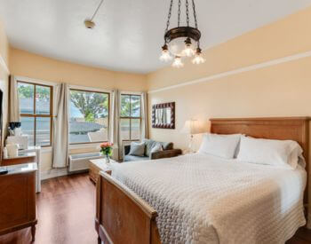 A bedroom with wood floors, yellow walls, a bed with a wood headboard and footboard, a red tapestry area rug, and a sitting area in front of bay window.