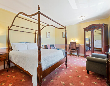 A bedroom with red and yellow carpeting, yellow walls, a four poster canopy bed, an antique mirrored wardrobe, a plush armchair, and writing desk.