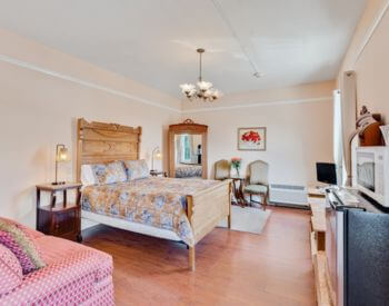 A bedroom with peach colored walls, dark wood floors, a bed with white bedding, a mirrored vanity on one wall, and a red loveseat along another wall.