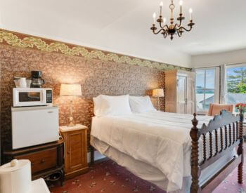 a bedroom with red floral carpeting, red tapestry wallpaper, an antique bed with wood spiral headboard and footboard, nightstands with lamps on either side of the bed, and a sitting area in front of a bank of windows.